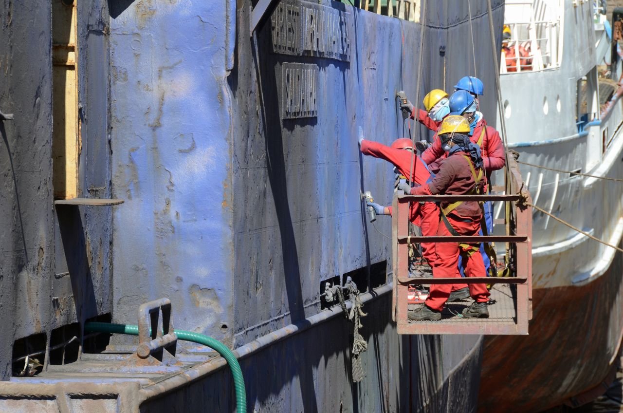 Workers in safety gear repairing a ship's hull at a busy shipyard.